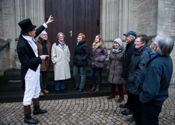 Ein Stadtführer erklärt einer Besucher*innen-Gruppe vor dem Willibrordi-Dom den Eingangsbereich des historischen Gebäudes.